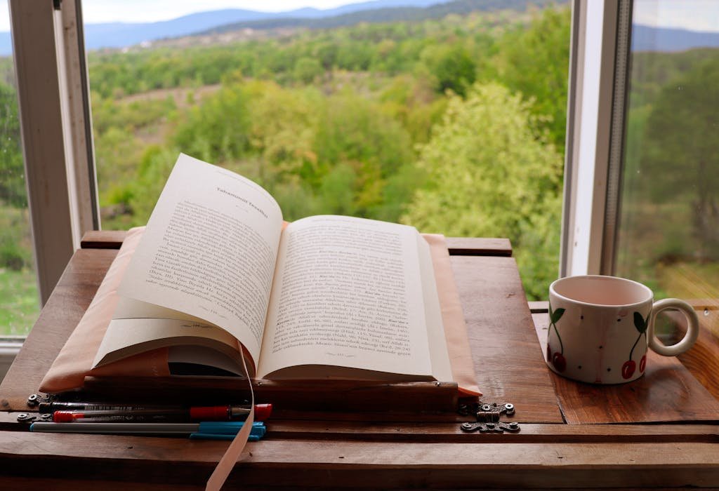 An open book and a cherry-patterned coffee mug on a table by a scenic window view.